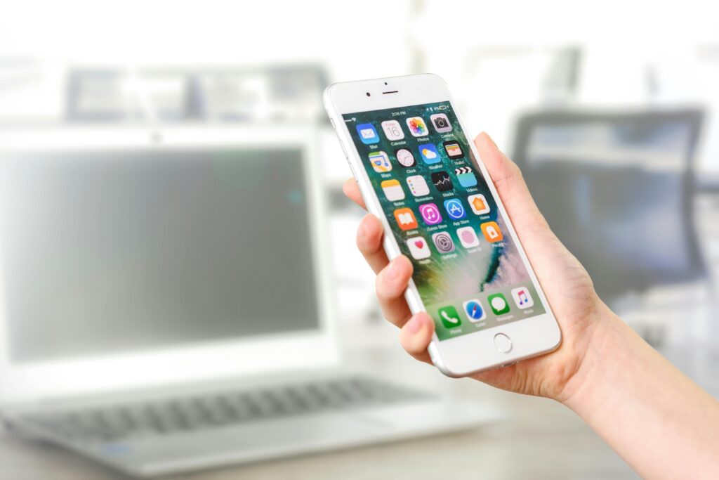 pexels-photo-887751-887751-1 Close-up view of a smartphone displaying apps, held by a hand, with a blurred laptop in the background.
